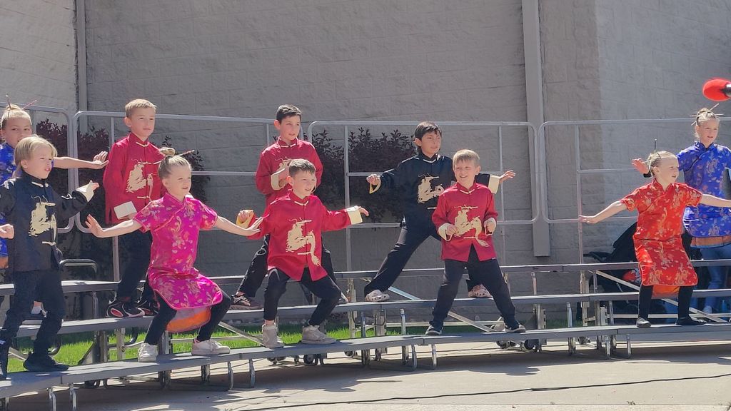 A group of elementary school students perform a dynamic dance on outdoor silver risers. The students are dressed in traditional Chinese tunics and dresses in vibrant colors like fuchsia, bright red, and black, many featuring gold dragon motifs. They are captured in a wide, athletic stance with their arms outstretched to the sides and knees slightly bent. The performance takes place on a bright, sunny day against the backdrop of a neutral-colored school building and dark red shrubbery.