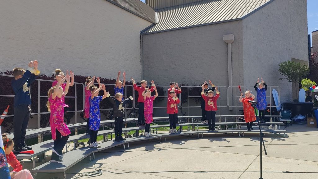 wide-angle outdoor shot of elementary students performing on a curved set of choral risers. The students are dressed in bright silk traditional Chinese costumes, including vibrant pink, blue, and red qipaos and tunics. Every student has their arms raised toward the sky with their hands open in a coordinated performance move. The stage is set on a paved area next to a large school building under a bright, clear blue sky. A microphone stand is visible in the foreground.