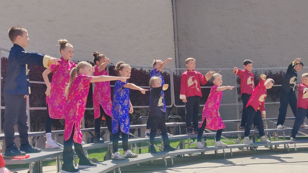 A group of elementary students perform a synchronized dance on outdoor choral risers. The students are wearing traditional Chinese attire, including bright pink, royal blue, and red qipaos and tunics with gold dragon patterns. Most of the students are captured mid-motion, pointing forward with one arm in a coordinated dance move. They are performing outside in front of a light-colored building wall on a clear, sunny day.
