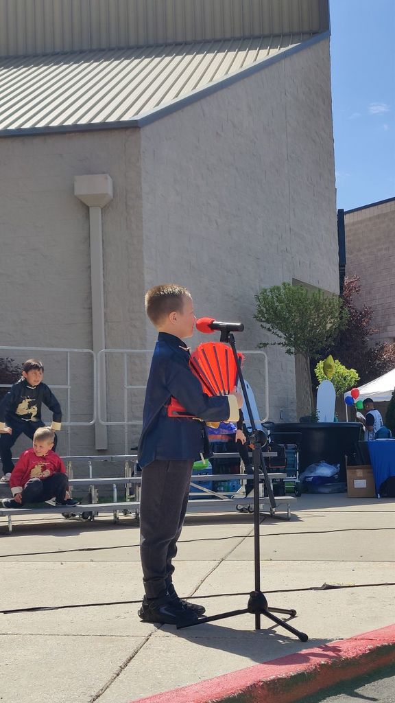 A young boy stands center stage at an outdoor microphone during a school performance. He is wearing a dark navy blue traditional Chinese tunic with gold trim and grey trousers. He is holding a folded red fan close to his chest while speaking or singing into a microphone with a red windscreen. In the background, two other students in traditional costumes are sitting on stage risers, and the school building is visible under a bright blue sky.