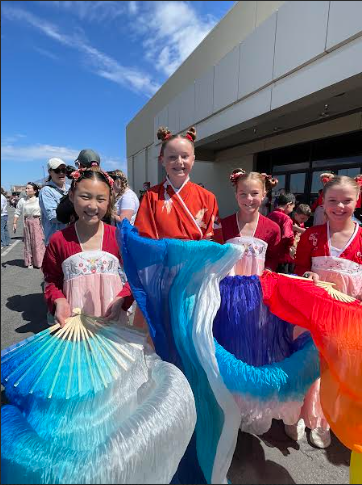 Four smiling young girls stand together outdoors, dressed in traditional-style Chinese dance costumes. Three of the girls wear pink and red dresses with embroidered floral bodices, while one wears a bright red robe. They have their hair styled in decorative buns with red accessories. Each girl holds a large, vibrant silk folding fan; the fans feature flowing gradients of blue, white, purple, and orange fabric. The sun is shining, and other people and a building are visible in the background.
