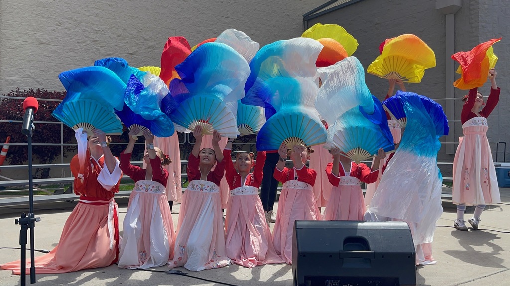A large group photo of approximately 20 elementary students and their teacher posing outdoors in front of a school building. The students are dressed in vibrant traditional Chinese performance costumes. In the front row, five boys in white shirts and red pants kneel while holding long red poles. Behind them, a line of girls stands holding large, flowing silk fans in a rainbow of colors including yellow, orange, blue, and purple. One student in the back holds two white prop swords crossed in the air. Everyone is smiling for the camera on a bright, sunny day.