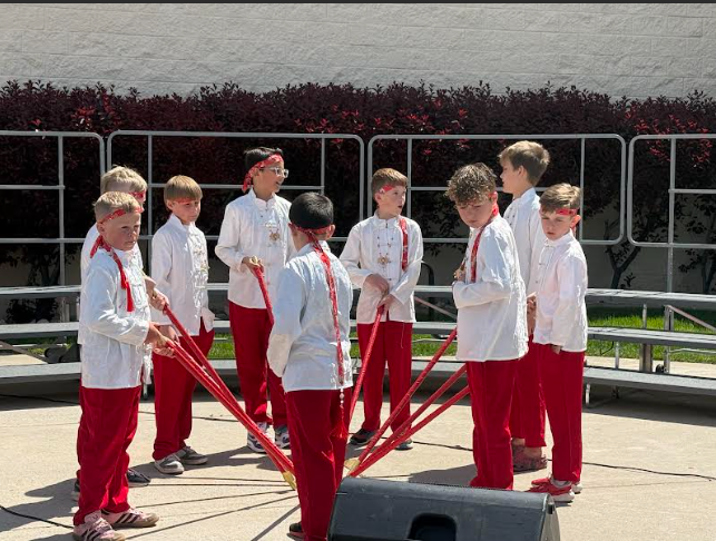 A group of about nine young boys performing an outdoor dance on a paved stage. They are dressed in matching costumes consisting of white long-sleeved button-down shirts with ornate detailing, bright red trousers, and red bandanas tied around their foreheads. Several boys are holding long red poles that meet in the center of their circle, creating a star-like pattern. They are standing in front of metal choral risers and a hedge of dark red bushes under bright, sunny skies.