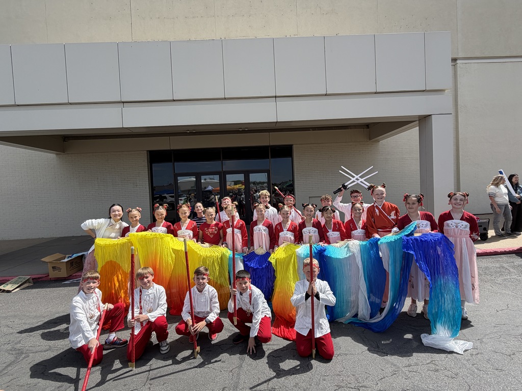 A large group photo of approximately 20 elementary students and their teacher posing outdoors in front of a school building. The students are dressed in vibrant traditional Chinese performance costumes. In the front row, five boys in white shirts and red pants kneel while holding long red poles. Behind them, a line of girls stands holding large, flowing silk fans in a rainbow of colors including yellow, orange, blue, and purple. One student in the back holds two white prop swords crossed in the air. Everyone is smiling for the camera on a bright, sunny day.