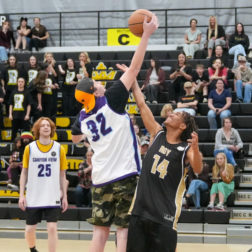 A jump ball or rebound situation at the hoop. A Canyon View player (number 32) and a Roy High player (number 14) both reach high for the basketball against the backdrop of a crowded gymnasium bleacher section.