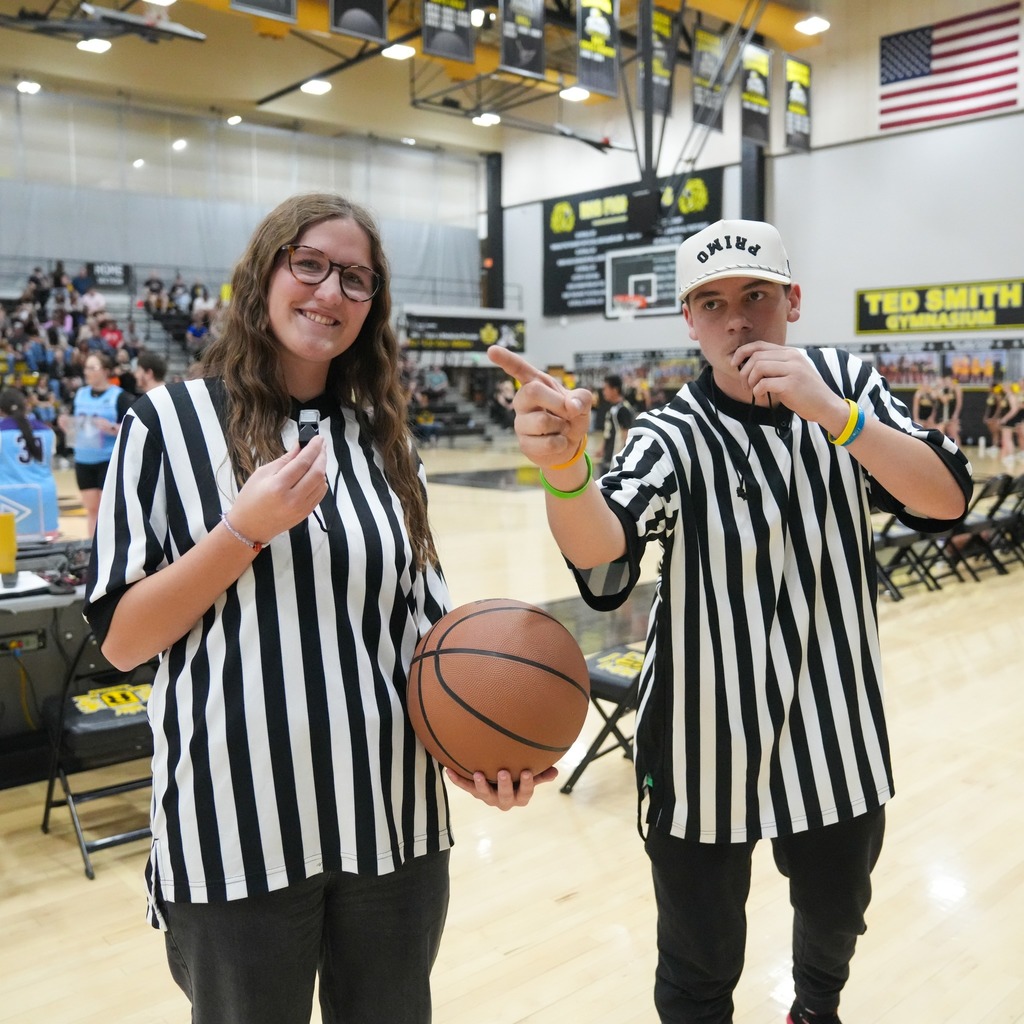 Two students acting as referees stand on the court wearing traditional black-and-white striped official shirts. One holds a basketball while the other points toward the court and blows a whistle.