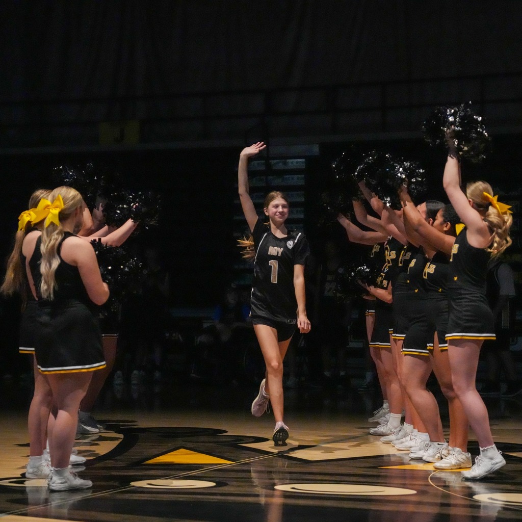 A spotlight shines on a Roy High athlete wearing jersey number 1 as she runs through a tunnel formed by the cheerleading squad, waving her hand to the cheering crowd during the starting lineup introductions.