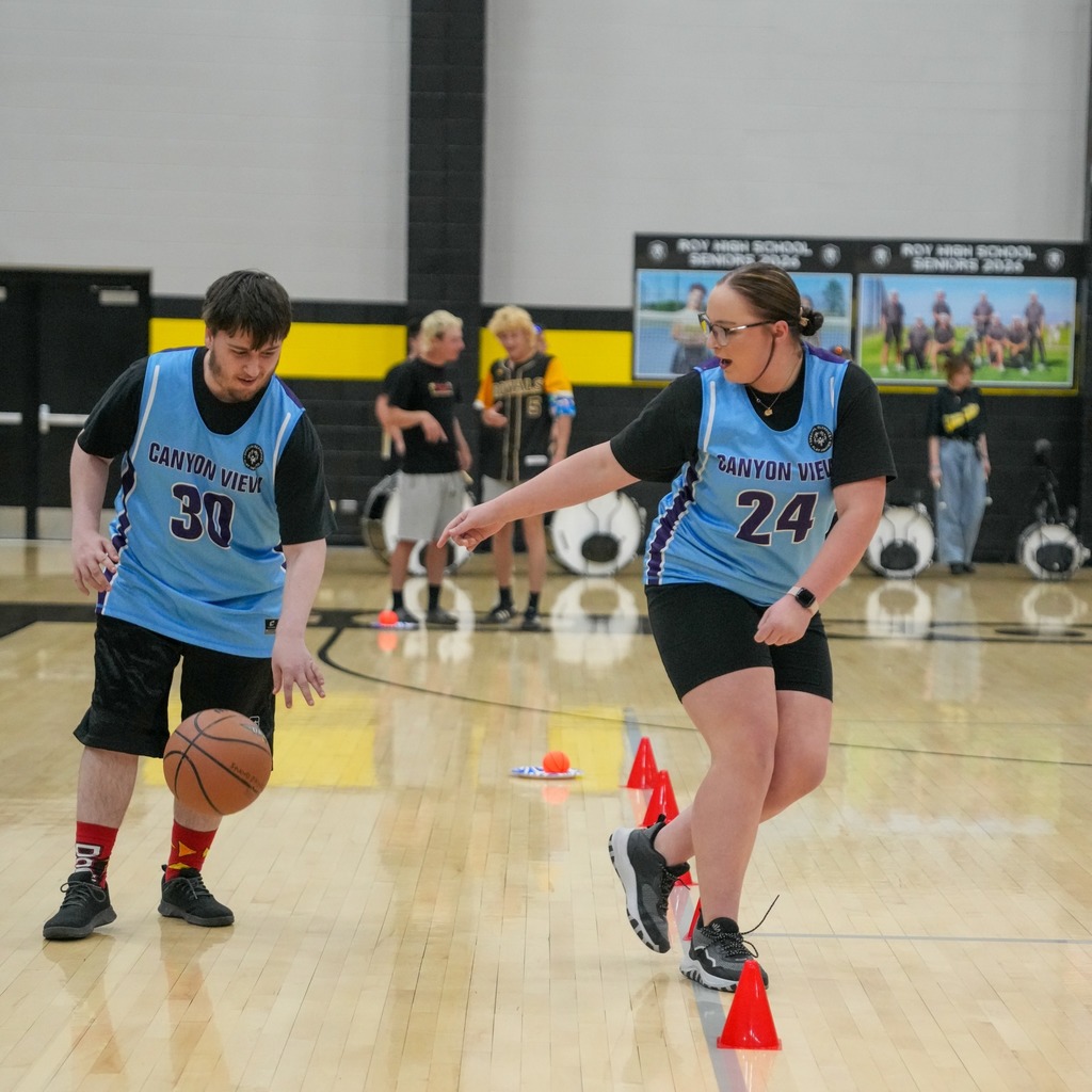 Two Canyon View students in light blue jerseys practice basketball drills on the Roy High gym floor. A male student (number 30) dribbles the ball while a female student (number 24) points toward a series of orange cones used for a skills course.