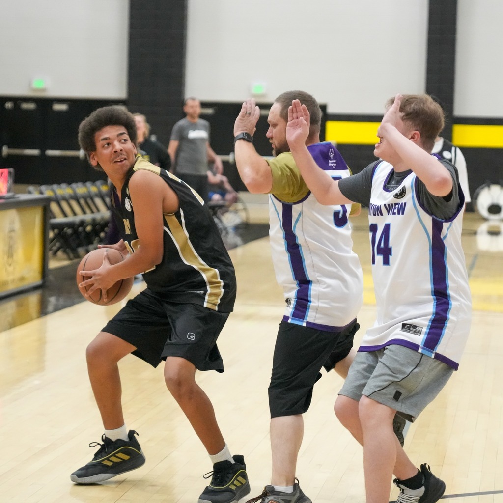 A Roy High player in a black jersey (number 12) looks for an open pass while being guarded by two Canyon View players in white jerseys who have their hands up in a defensive stance.