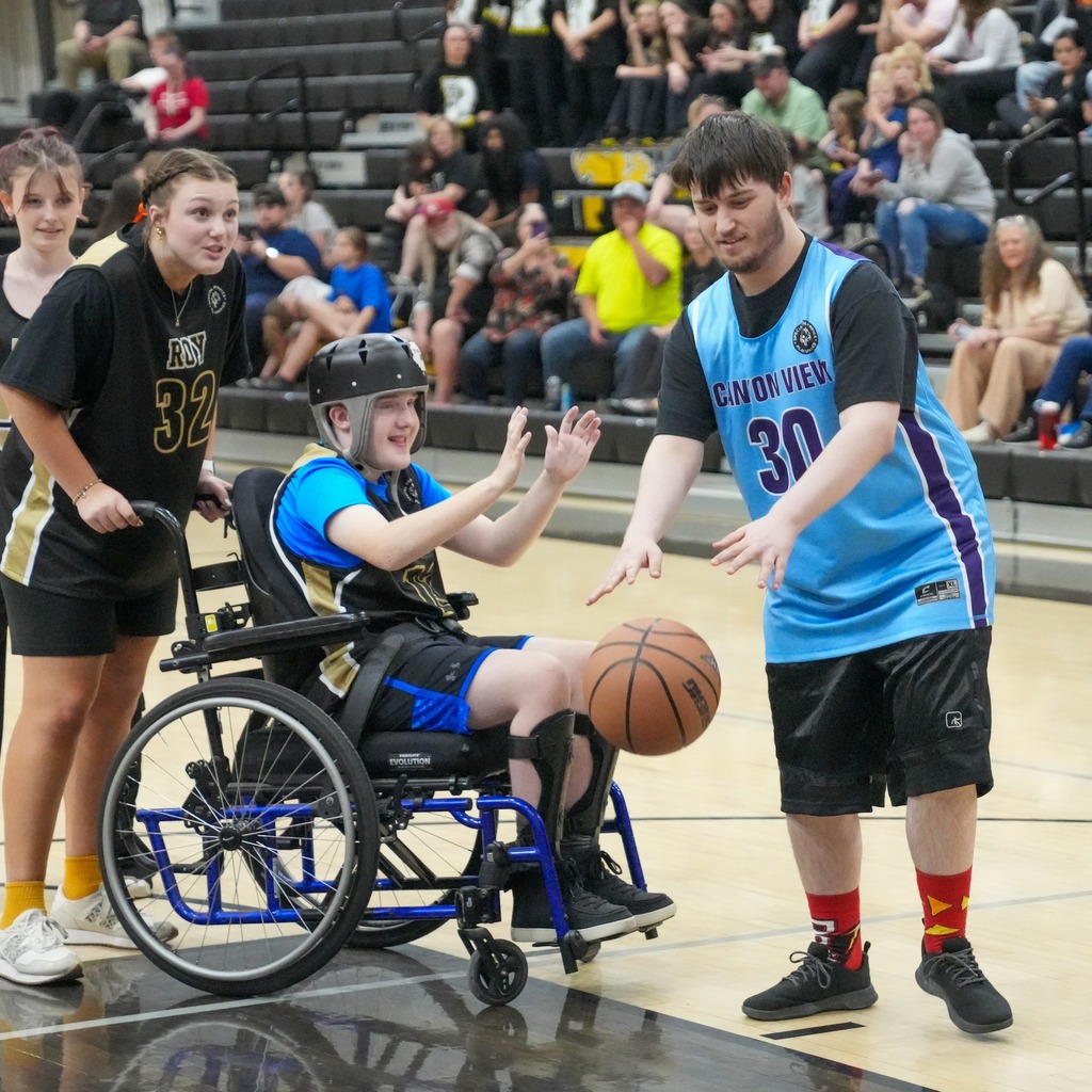 A heartwarming moment of sportsmanship on the court. A Canyon View student (number 30) smiles as he bounces the ball toward a teammate in a wheelchair who is wearing a helmet and clapping with excitement.