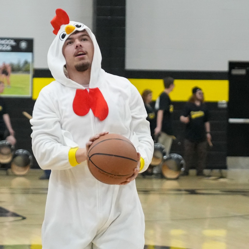 A student dressed in a full-body white chicken costume participates in the basketball festivities, holding a basketball and looking up toward the hoop with focus.