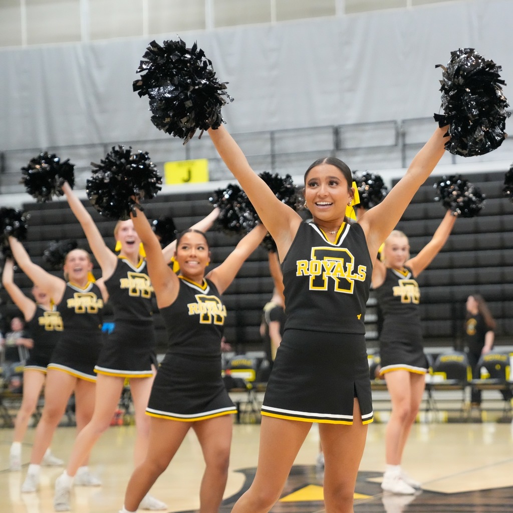 The Roy High Royals cheer squad performs a routine in the gymnasium. The cheerleaders are wearing black and gold uniforms and holding large black pom-poms high in the air with big smiles.