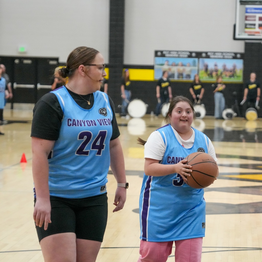 Two female Canyon View athletes in blue jerseys walk down the court together. The student in the foreground (number 5) holds the basketball with a bright, joyful expression while her teammate (number 24) walks alongside her.