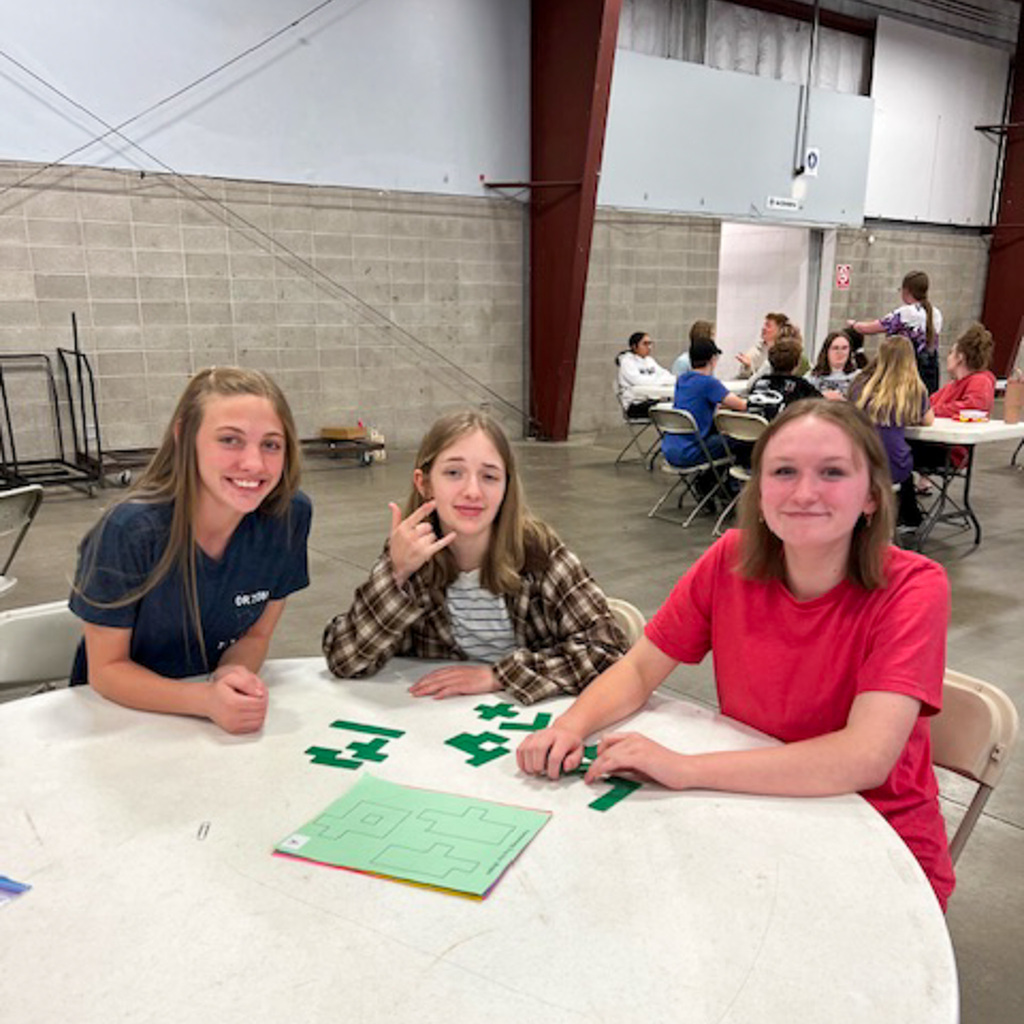 Three female students smile at the camera while working on a green geometric puzzle task at a round table in the fairgrounds exhibition hall.
