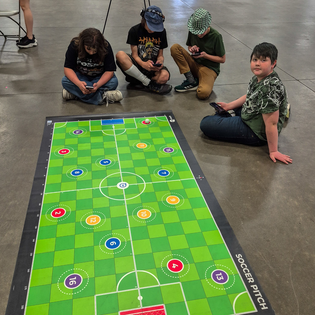 Four students sit on the floor around a large "Soccer Pitch" mat, using their smartphones to program and control Sphero Robots for a STEM challenge.