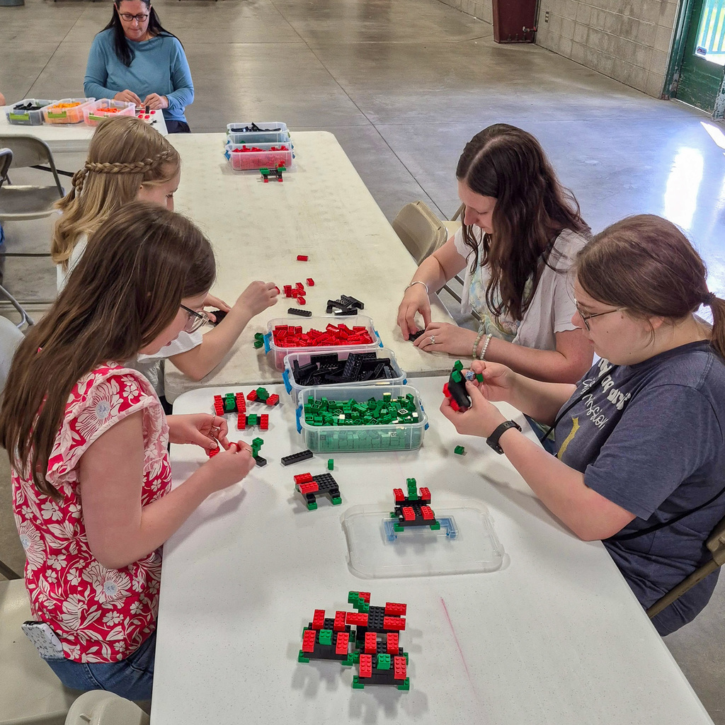 Four students sit at a long table focused on building intricate models using green, red, and black plastic building bricks during the MESA competition.