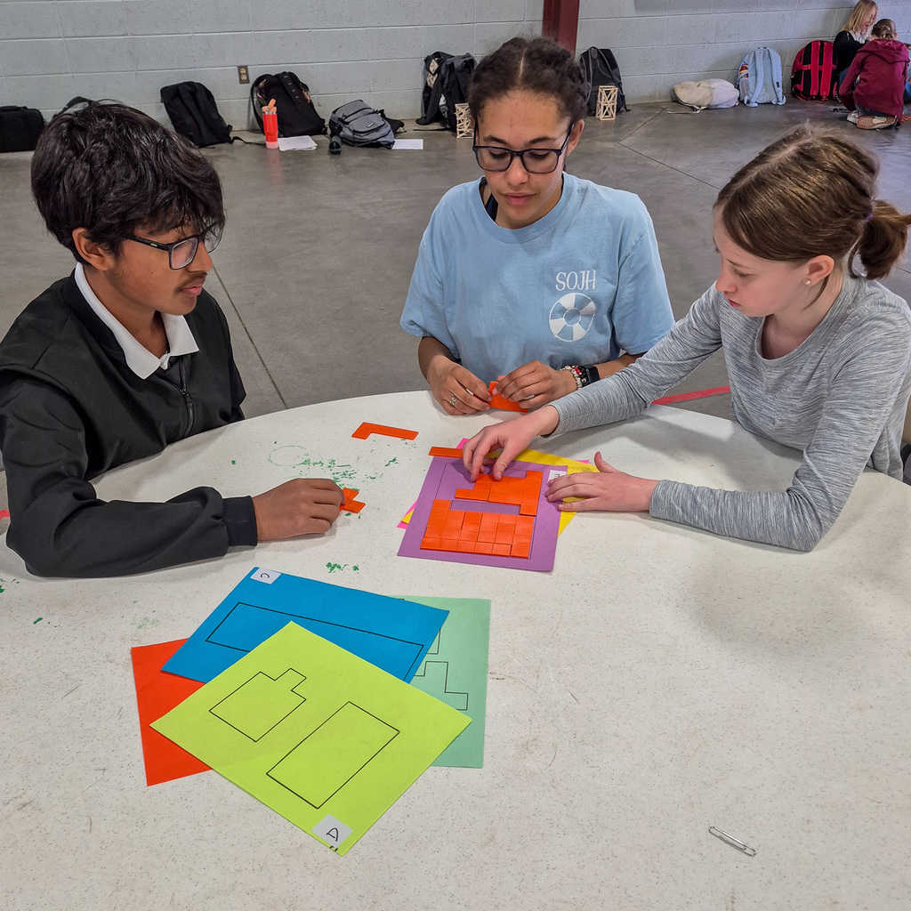 Three students sit at a round table working collaboratively on a Pentominoes puzzle, carefully arranging orange geometric shapes on a purple template.
