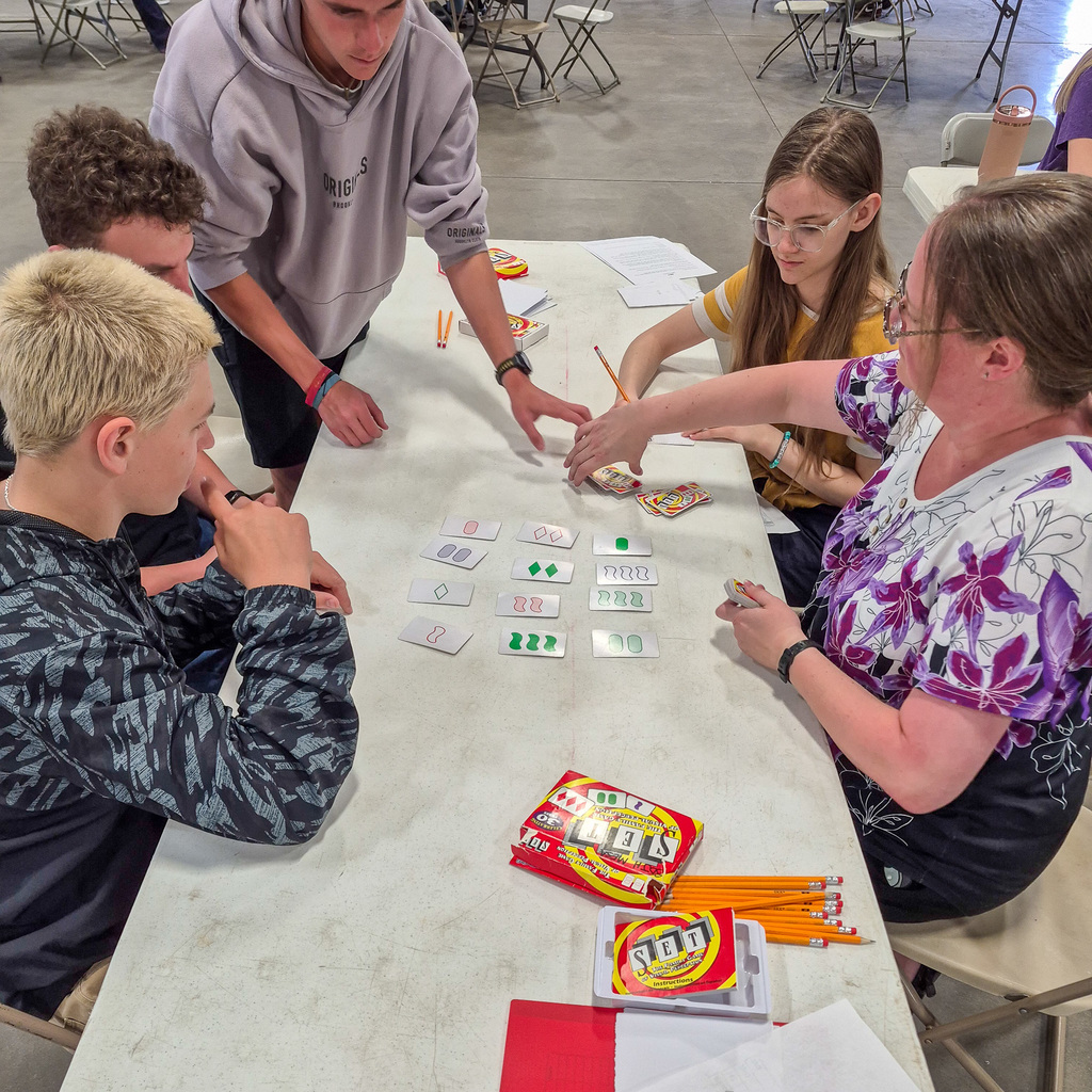 A group of students and an advisor gathered around a table playing the card game "Set," focusing on identifying patterns among the cards laid out in a grid.
