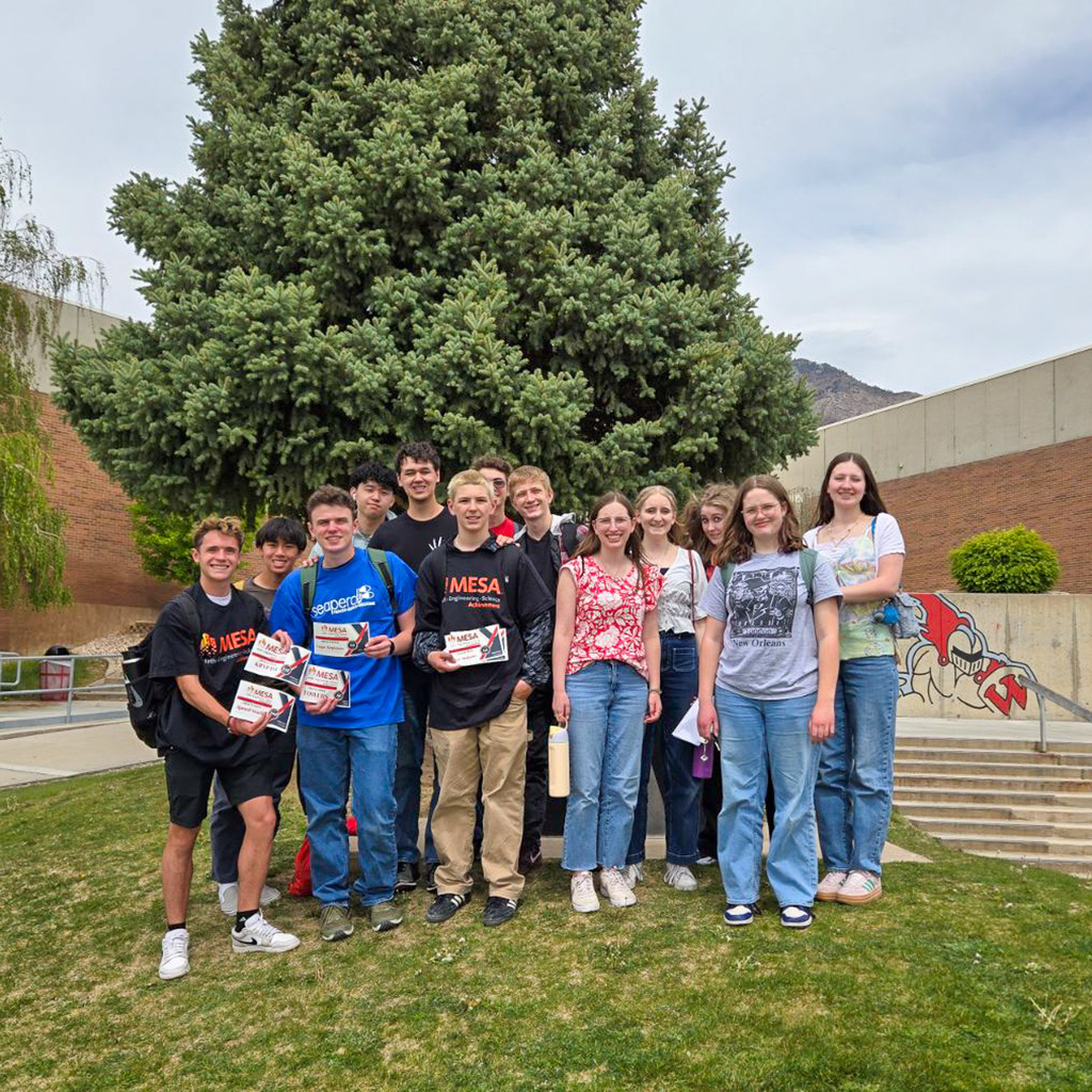 large group of high school students pose together outside in front of a large evergreen tree, proudly holding their MESA award certificates.