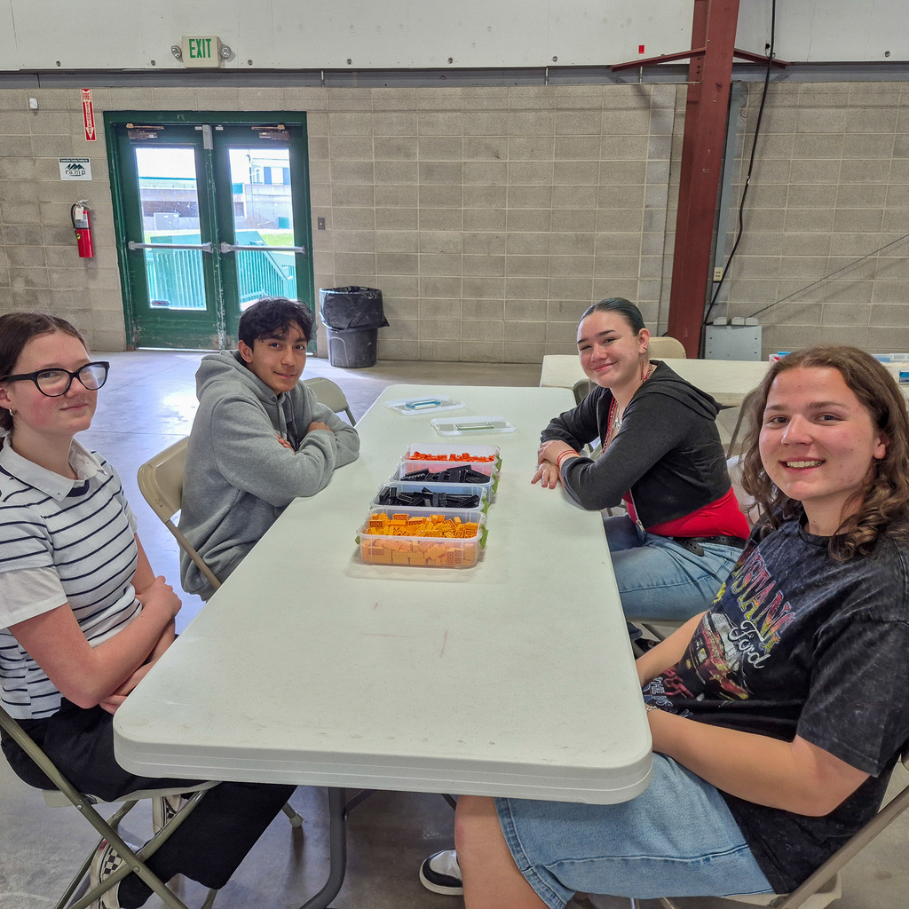 Four smiling students sit together at a table with containers of building blocks, ready to begin their next MESA engineering challenge.