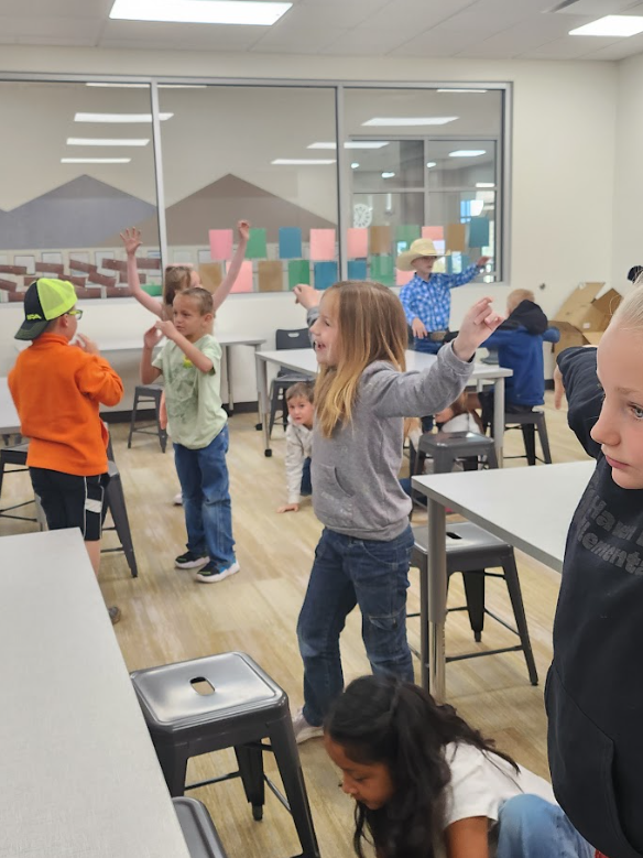 A group of children in a classroom are standing up and moving their arms, appearing to mimic animal movements or stretch. A boy in a cowboy hat and blue plaid shirt is visible in the background, and another child wears a bright orange sweatshirt and neon yellow hat.