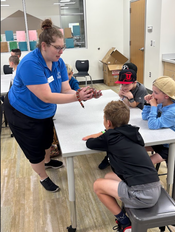 A presenter named Kellie in a blue aquarium polo shirt leans over a table to show a small, orange, black, and white banded snake to three young boys. A box in the background is labeled "GLASS FRAGILE."