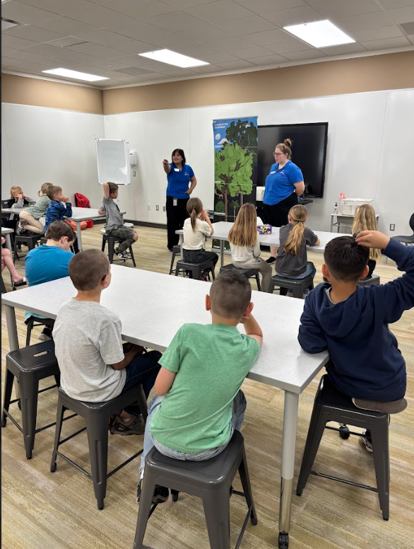 A wide shot of a classroom setting where two presenters in blue "Loveland Living Planet Aquarium" shirts stand at the front. A banner reads "Layers of the Rainforest" with sections labeled "Emergent," "Canopy," "Understory," and "Forest Floor." Students sit on stools at white tables listening to the presentation.