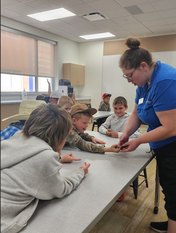A presenter in a blue shirt and glasses holds a small dark creature for a group of students at a long table. One boy in a camouflage jacket reaches out to touch it while other students watch with expressions of curiosity and excitement.