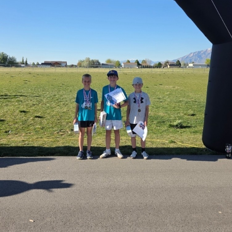Three young boys stand under a large, black inflatable finish line arch on a paved path. Each boy is wearing a teal or grey shirt and a medal around his neck. The two boys on the right hold paper certificates and boxes. The background shows a grassy field and distant snow-capped mountains.