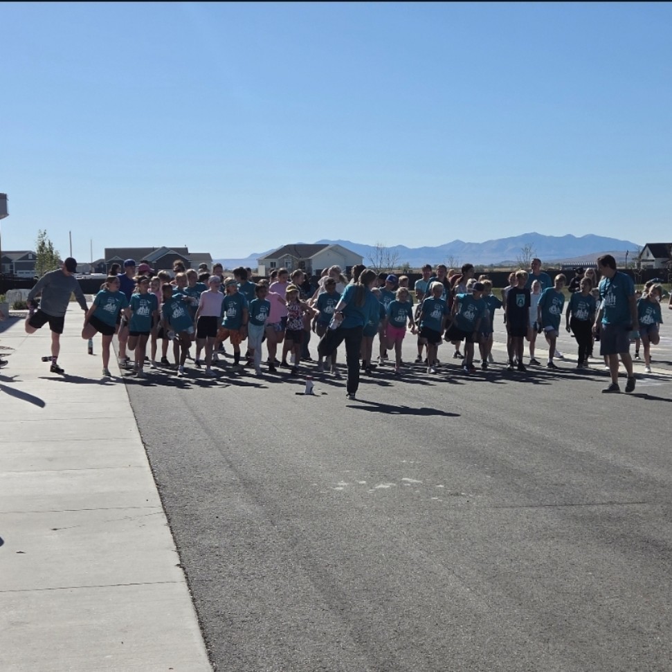 A large group of children and a few adults in teal shirts stand on an asphalt path outdoors, performing a standing quad stretch before a race. In the background, residential houses and a mountain range are visible under a clear blue sky.