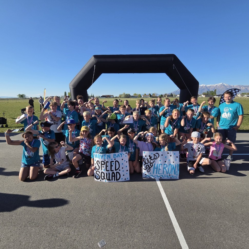 A large group of children in teal "Speed Squad" shirts pose for a group photo in front of a black inflatable arch. Many are flexing their muscles. Two children in the front hold signs: one says "HAVEN BAY SPEED SQUAD" and the other says "LET'S GO HERONS." A man stands on the far right holding a checkered flag.