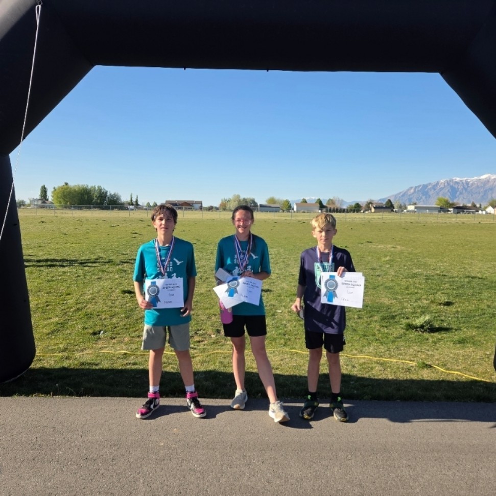 Three young boys in teal shirts stand under a black inflatable arch wearing medals. They are looking down at the white certificates they are holding. The scene is set against a backdrop of a green field and a mountain range.