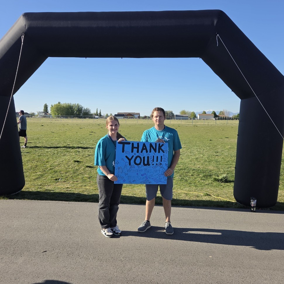 A young woman and a man stand under a black inflatable arch holding a large blue handmade sign that says "THANK YOU!!!" in bold black letters. Smaller text on the sign includes names like "Mr. Weston," "Mrs. Martin," and "Ms. Sam." They are both wearing teal shirts and standing on a paved path.