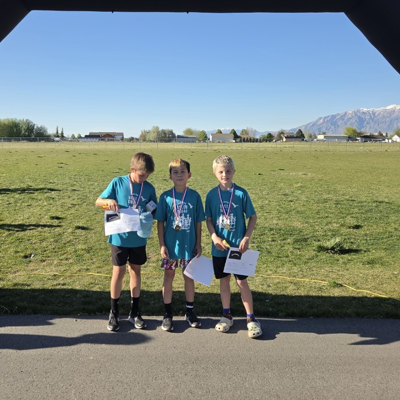 Three young boys in teal shirts stand under a black inflatable arch wearing medals. They are looking down at the white certificates they are holding. The scene is set against a backdrop of a green field and a mountain range.