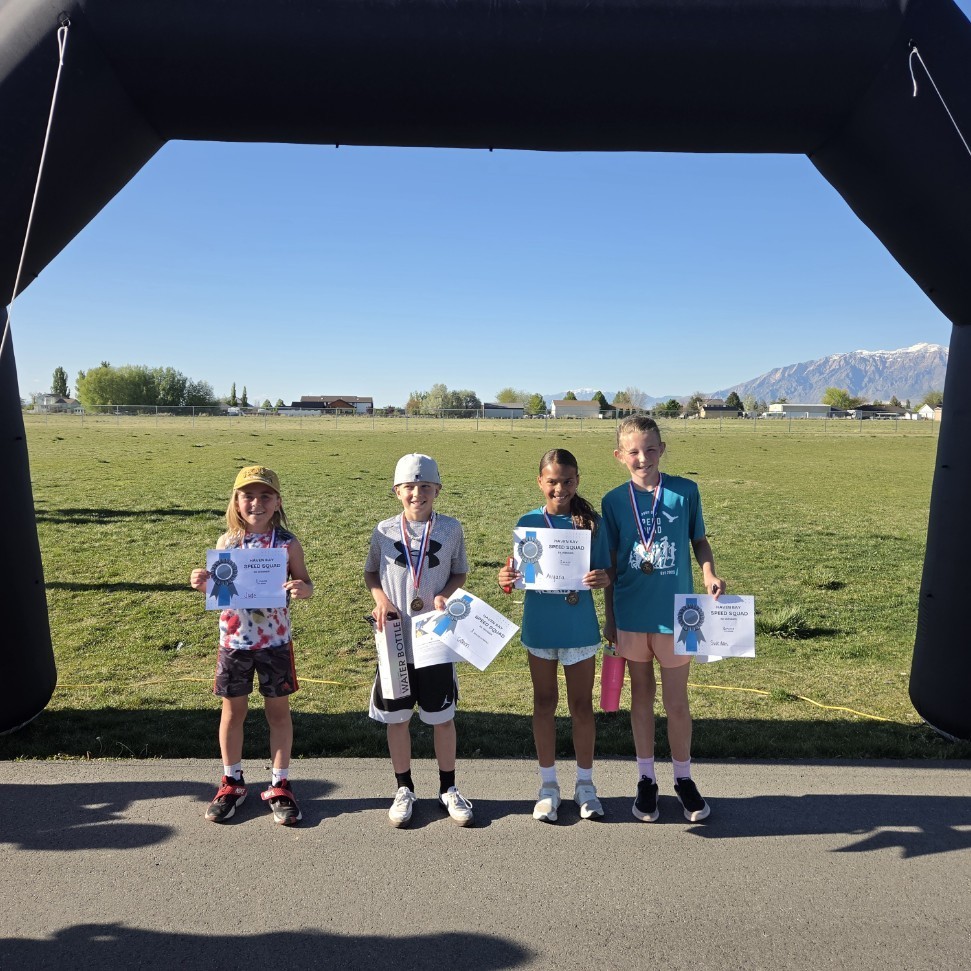 Four children stand in a line under a black inflatable archway. They are all wearing medals and holding white award certificates that say "Haven Bay Speed Squad." The group includes three girls and one boy, all dressed in athletic wear.