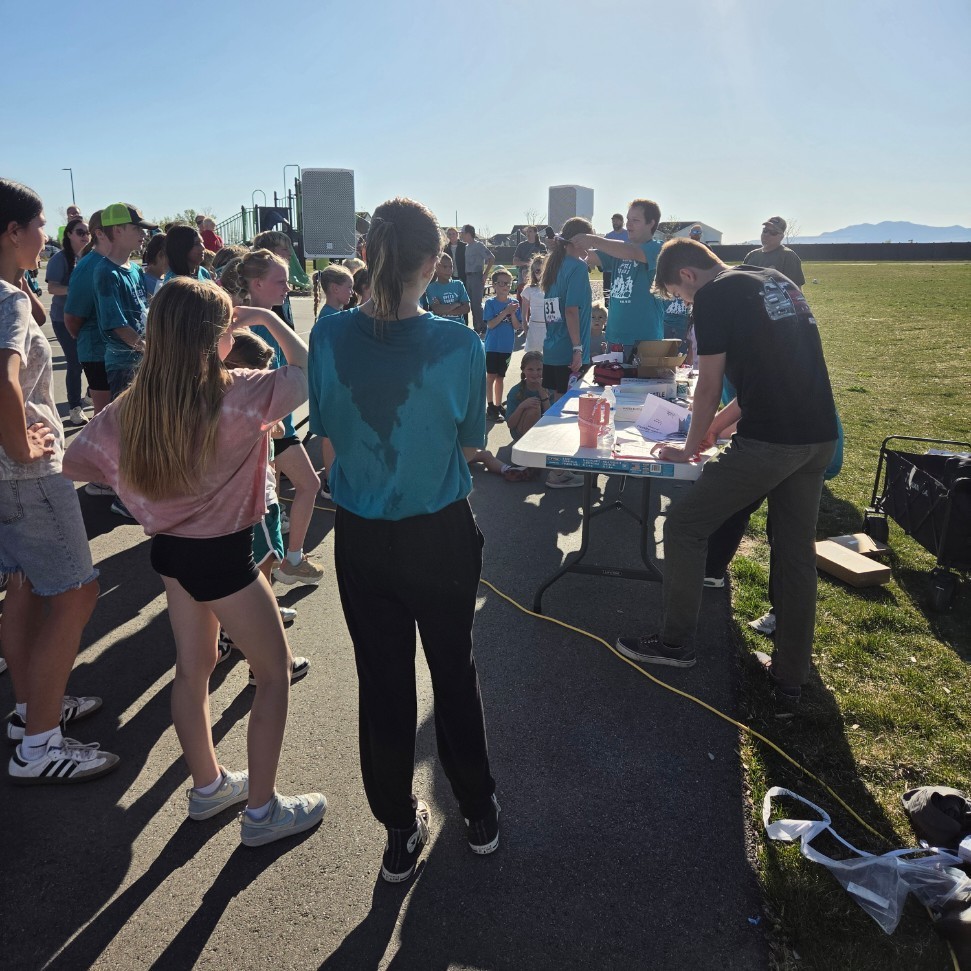 A view from behind two large white PA speakers on stands, looking toward a crowd of children in teal shirts gathered in a parking lot. A woman in a red jumpsuit stands to the left, and a white table with a megaphone is on the right. The sun is shining brightly overhead