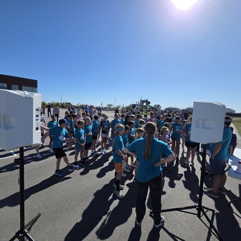 A view from behind two large white PA speakers on stands, looking toward a crowd of children in teal shirts gathered in a parking lot. A woman in a red jumpsuit stands to the left, and a white table with a megaphone is on the right. The sun is shining brightly overhead