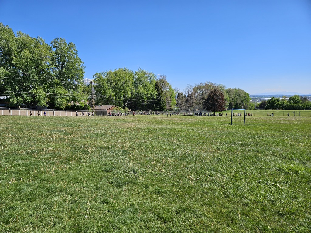 A wide shot of a lush green field at Lomond View Elementary under a clear blue sky, with many students running along the perimeter fence.