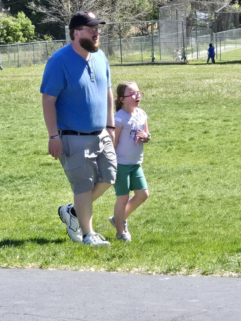 A bearded man in a blue shirt and a young girl with glasses walk together across the grass during the fun run.