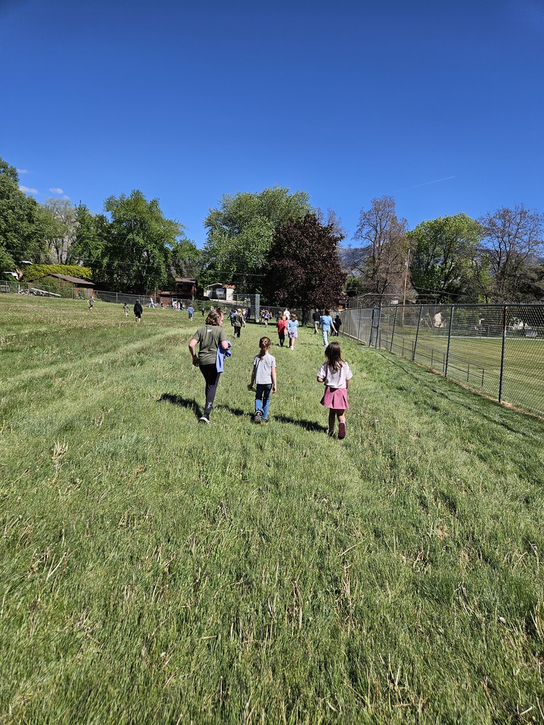 Three young girls walk across a grassy hill toward the finish line, with the school and mountains in the background.