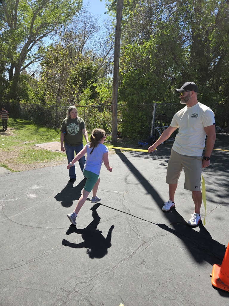  A young girl in a blue shirt crosses a finish line marked by yellow tape held by a volunteer in a white shirt and hat.
