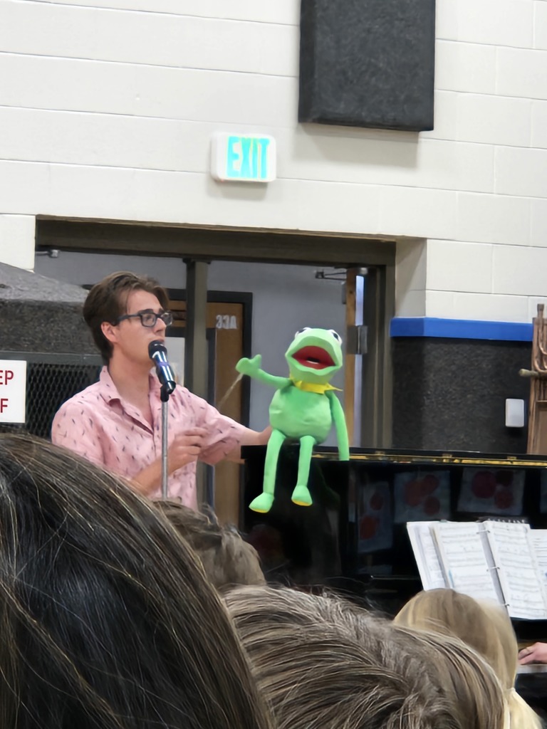 A performer in a pink shirt holds a Kermit the Frog puppet up to a microphone near a piano.