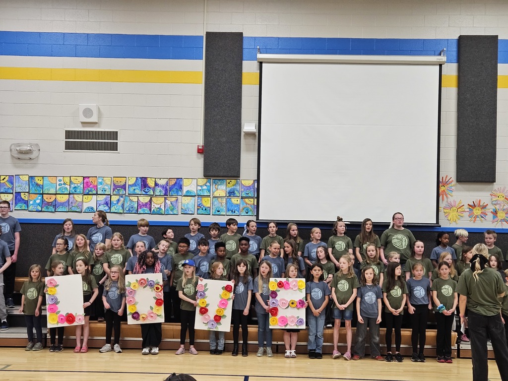 The school choir stands on risers holding large white signs that spell out "LOVE," decorated with colorful paper flowers.