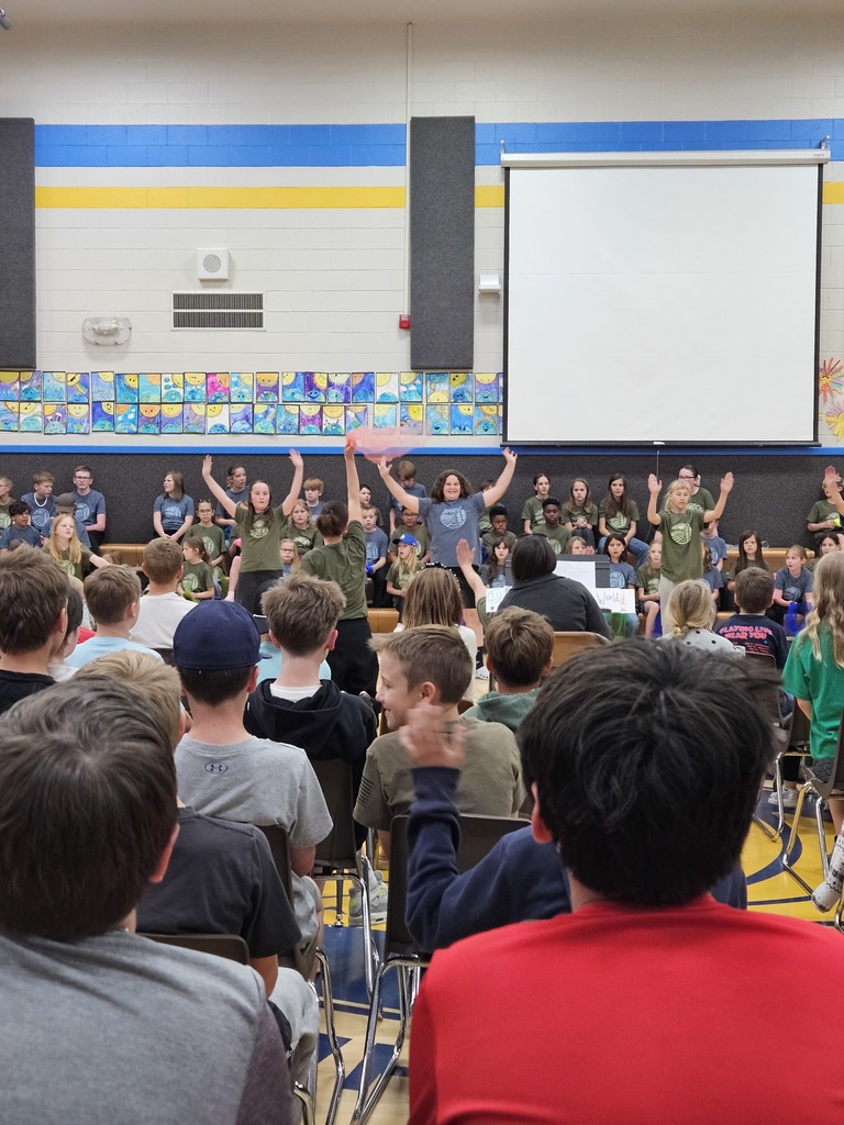 Students in green and blue shirts perform on a stage in the gym, some holding a pink scarf, while an audience of students watches from the foreground.