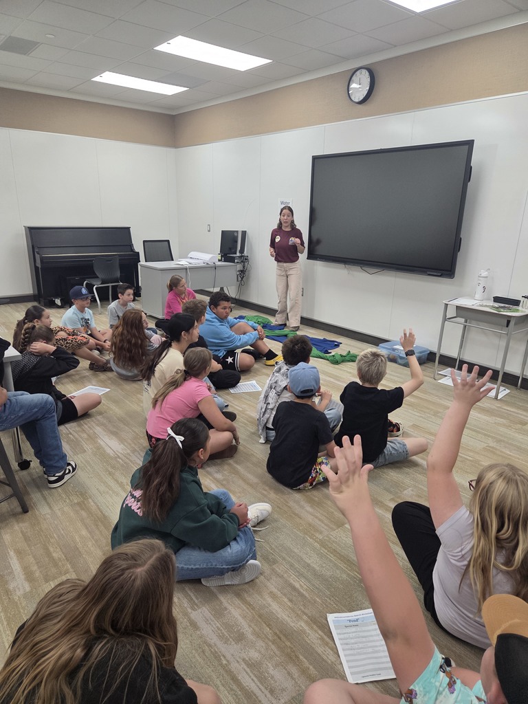 An instructor stands at the front of a classroom next to a large, blank digital display, addressing a group of about fifteen students sitting on the floor. Some students have their hands raised. A piano and a teacher's desk are in the background. A student in the foreground holds a Macroinvertebrate Search Worksheet for "Pond". A small sign on the wall behind the instructor reads Water.