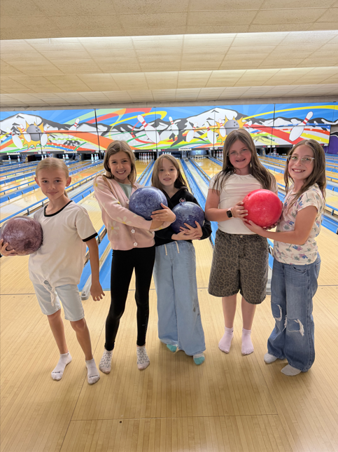 Five elementary-aged girls standing in a row on a bowling lane, hugging their bowling balls and smiling broadly for the camera.