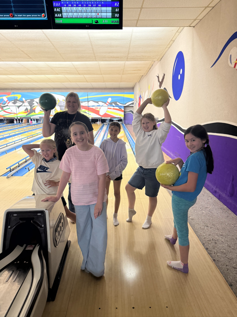 A group of five smiling elementary girls and one adult woman posing on a bowling lane. Two of the girls and the adult are playfully holding bowling balls over their heads or out to the side, while others pose near the ball return. A digital scoreboard is visible in the background showing their scores.