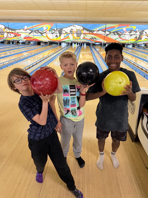 Three young boys standing on a bowling lane, smiling and holding their bowling balls (red, black, and yellow) up toward the camera.