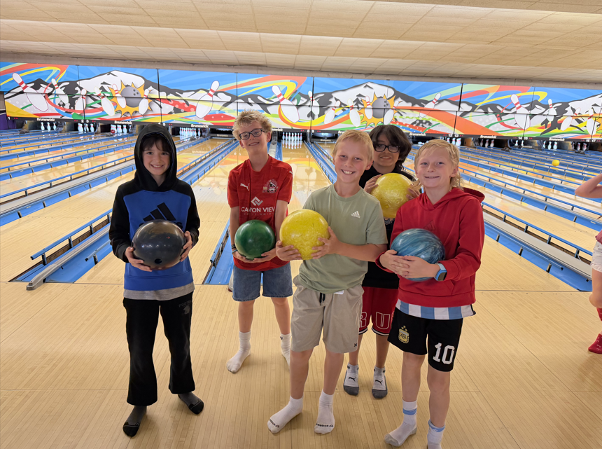 Five boys posing together at the end of a bowling lane, each holding a different colored bowling ball and wearing socks, ready to play.