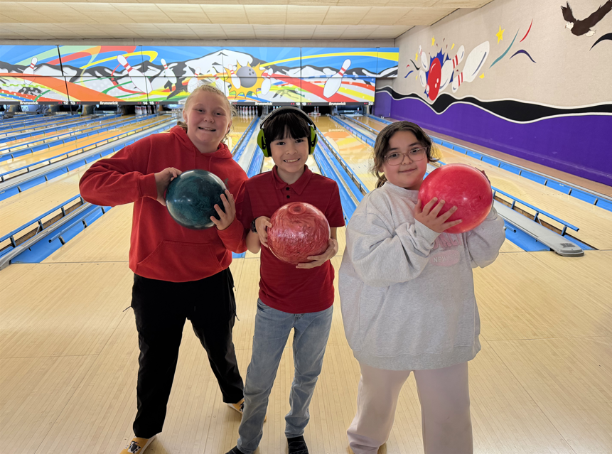 Two girls and one boy smiling for a photo at the bowling alley. The boy in the center is wearing bright green noise-canceling headphones and holding a red bowling ball.